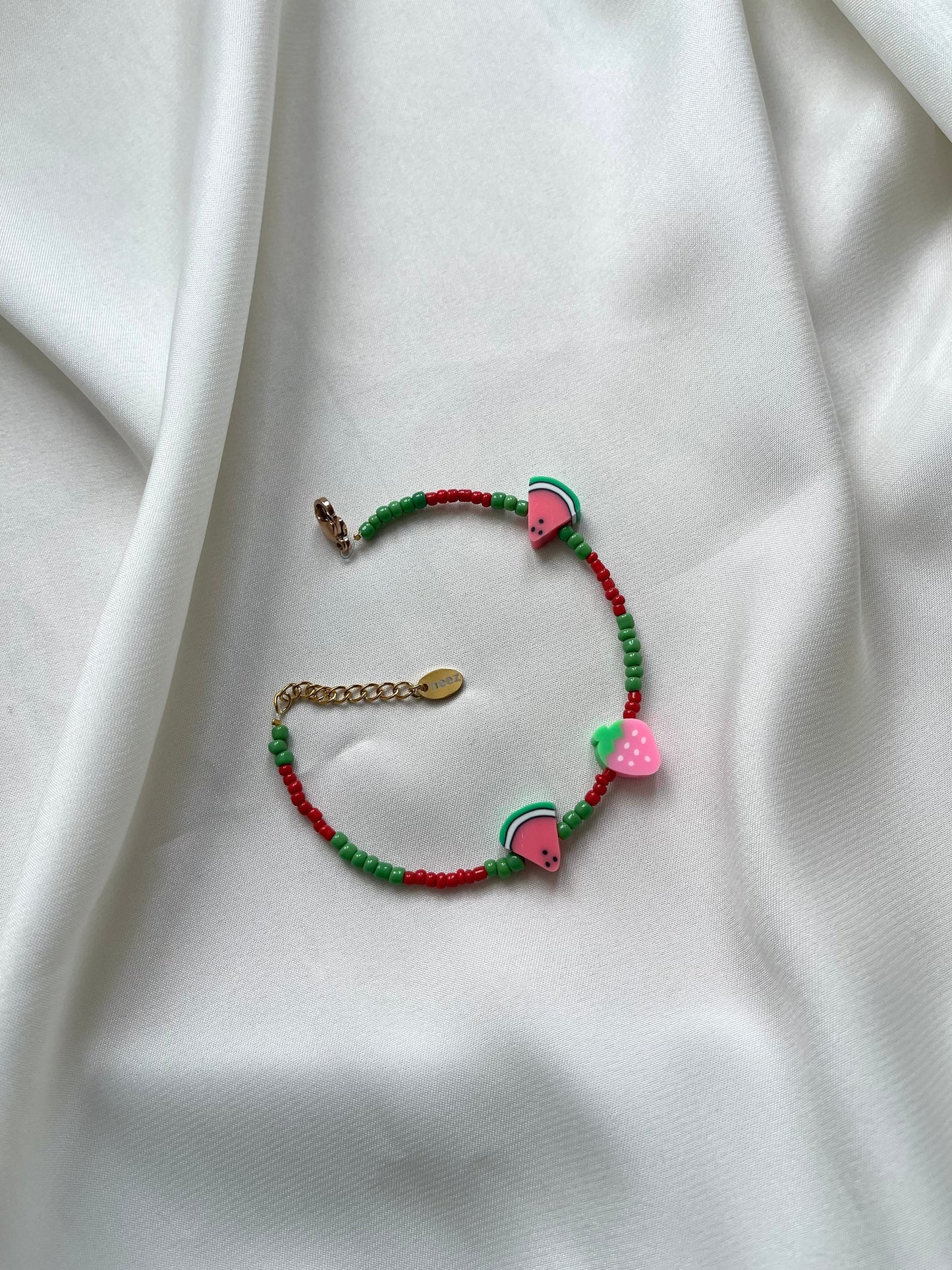 A multicolored bracelet with green, red, and pink beads, featuring a metallic clasp, displayed on a white fabric surface.
