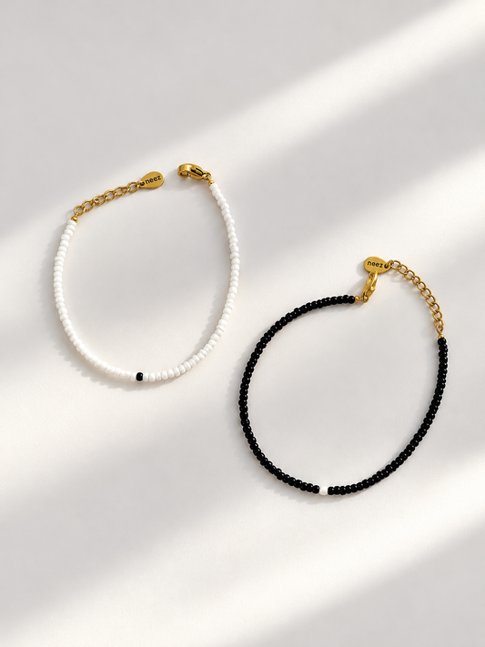 Two beaded bracelets, one white and one black, each featuring a gold chain extender and clasp, displayed on a light gray background with soft diagonal shadows.