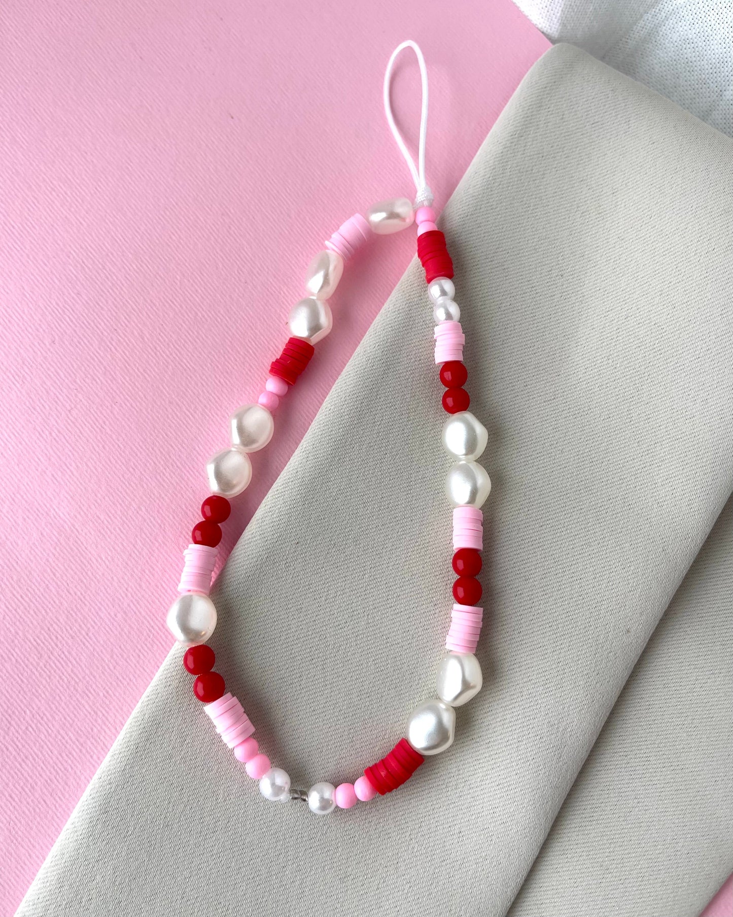 A multicolored phone strap with white, red, and pink beads, placed on a pink background next to a white napkin.