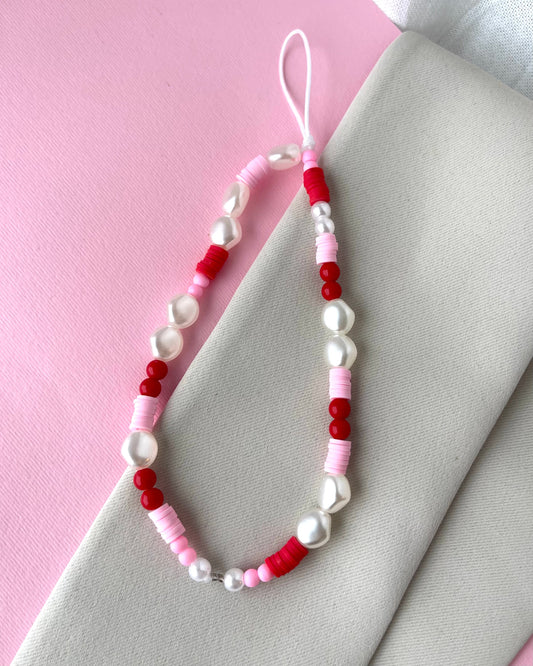 A multicolored phone strap with white, red, and pink beads, placed on a pink background next to a white napkin.