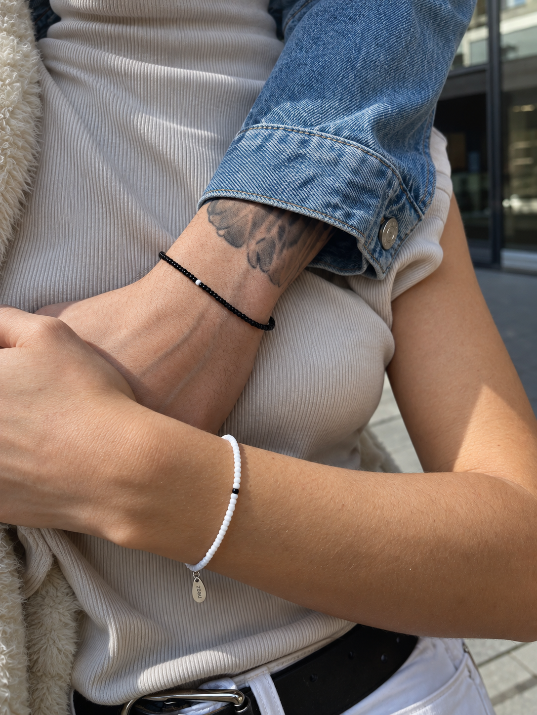 A close-up lifestyle shot of a person's forearm wearing a delicate white beaded bracelet with a small silver charm, alongside other bracelets on the other wrist.
